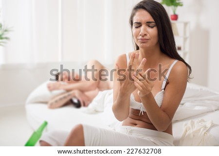 A young woman, just awakened, sitting on the bed with a sad expression on her face trying to take off the wedding ring from her finger, and next to her sleeping man hugging a bottle of drink.