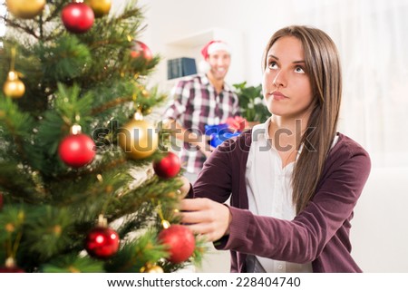 Young beautiful girl decorates a Christmas tree while her boyfriend holds a Christmas gift and he wants to surprise her.