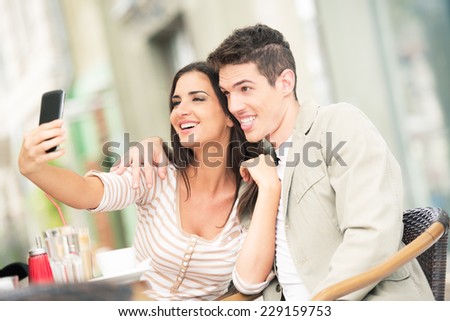 Young heterosexual couple sat embraced in a cafe, and a girl with her cell phone photographs of herself and her boyfriend.