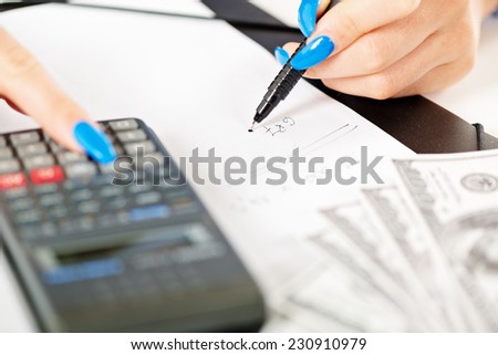 Close-up of calculator on white paper and women hands with long painted nails on the fingers that hold a pencil and write down the numbers on the paper next to a dollar bill.