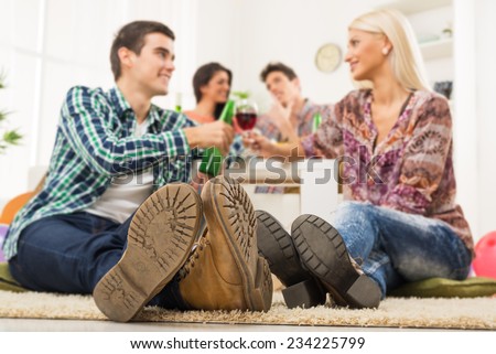 Young couple at house party with faces out of focus in the foreground are the soles of their shoes, sit on the floor, knocking the toasting glasses, in the background you can see another couple.