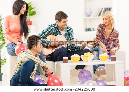 A small group of young people, friends at home party. A young man sitting on the couch between two beautiful girls, playing acoustic guitar and a guy sitting on the floor next to the stool