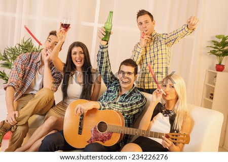 Group of young people at home party smiling looking at camera. A guy with a guitar and a girl next to him toasting with drinks, and other companies in the entertainment blowing in party whistle.
