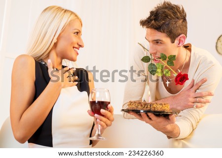 A young man with a rose in his mouth gives beautiful blond girl birthday cake, pleasantly surprised girl looking at him with a smile on her face.