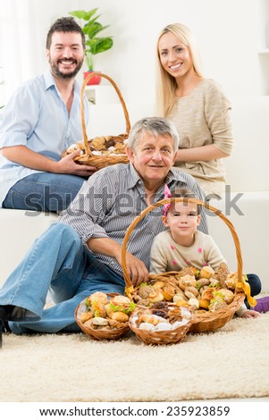 Senior man and a little girl are sitting on the living room floor. In front of them is woven basket with pastries, and behind them young parents are sitting on the couch and hold a basket of pastries.