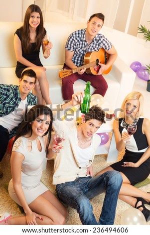A small group of young people at home party, photographed from above, looking at camera smiling, enjoying the time together with music, drinks and snacks.