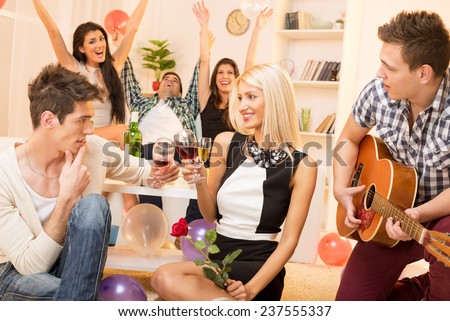 A young guy is courting the pretty girl at home party, while their friend plays acoustic guitar, and in the background the rest of society with arms raised celebrated their relationship.
