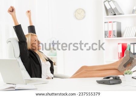 Young pretty blonde woman in the office sitting in office chair, hands up and with her feet up on a desk. In the background you can see the shelves with binders.
