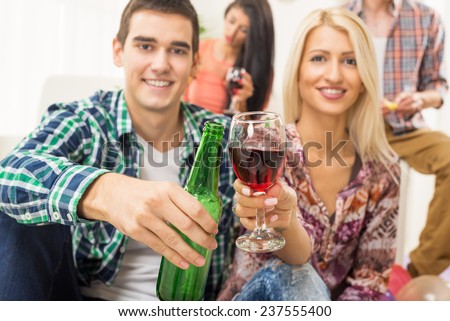 Young couple at home party with a smile on their faces out of focus, toasting with drinks that are in the foreground. In the background is another young couple.