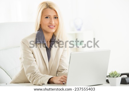 A young blonde businesswoman, elegantly dressed sitting at a desk in front of laptop in her office with a smile on her face looking at the camera.