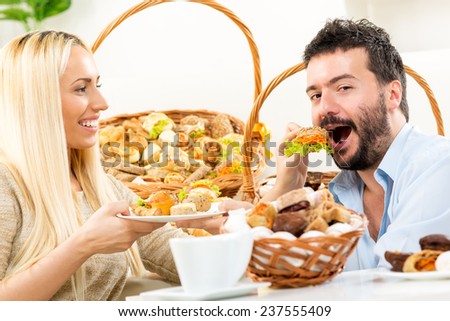 Beautiful blonde girl feeds her man with a pastry. In the background you can see woven baskets with bakery products.