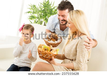 Young parents hold in their hands woven basket with delicious salty and sweet pastries. Beside them sit on the couch their little daughter eating pastries and holding thumb-up.