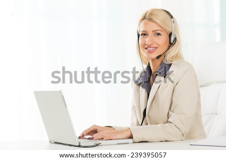 Young beautiful blonde girl, elegantly dressed, sitting at an office desk in front of laptop with a headset on her head. With a smile looking at the camera.