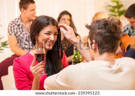 The young man at the house party is courting the pretty brunette caressing her hair, and in the background you can see a small group of young people having fun at a party.