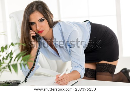 A young pretty woman in a short skirt, kneeling on a office chair, leaning on office desk, phoning and written in the planner.