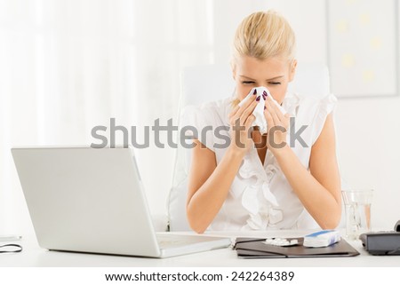 A young blonde business woman at her workplace at an office desk in front of laptop, shaking her nose into a handkerchief ready to take the pills.