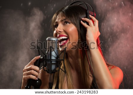 Portrait of a young woman singer with headphones in front of the microphone. Sing with mouth wide open and with an expression of happiness on her face.