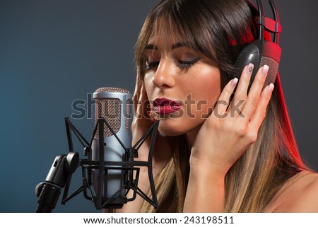 Beautiful young woman singer with the headphones standing in front of a microphone and sings with a slightly open mouth and eyes closed.