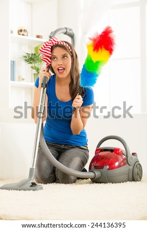 A young housewife kneeling beside the vacuum cleaner with duster in hand, satisfied with the good work cleaning up the house.