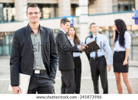 Young businessman with a laptop standing in front of office building separated from the rest of the business team. With a smile looking at the camera.