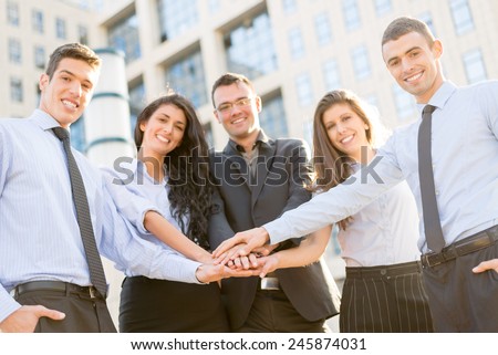 Small group of young business people standing in front of the company with folded hands, motivating for new business wins. With a smile, looking at the camera.