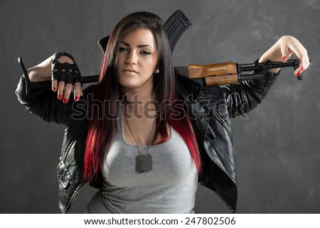 Attractive young woman with an automatic rifle standing with hands leaning on the rifle that was put behind the neck. With a dangerous expression on her face looking at the camera.