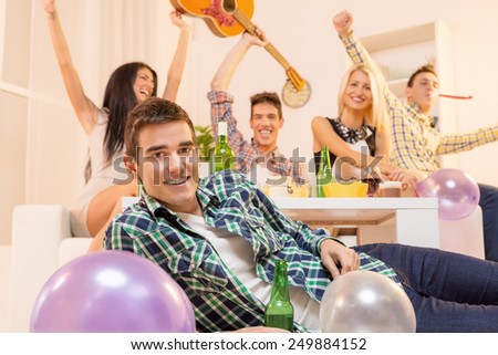 Young guy at house party, lying on the floor, smiling, looking at the camera in the background you can see his friends sitting on the couch happy with their hands raised.