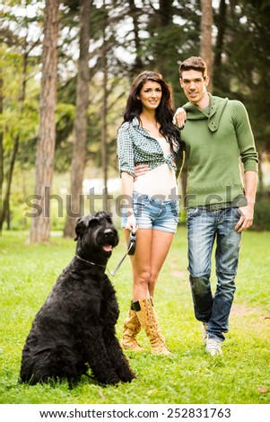 Young handsome heterosexual couple with a dog, a black giant schnauzer,  walking through the park.