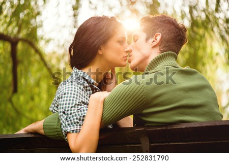 Young heterosexual couple photographed from behind while sitting on a park bench, kiss, illuminated by the rays of the spring sun.