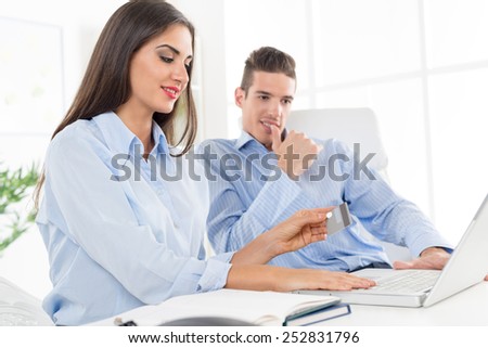 Two young smiling business people, man and woman sitting in office at an office desk in front of a laptop. Woman holding a credit card.