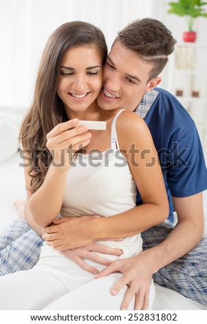 Young woman sitting on the bed with a smile on her face looking at pregnancy test, behind her is a young guy in pajamas, looking at pregnancy test with a smile and hugs girl.