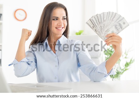 Young smiling businesswoman sitting in the office, in one hand holding dollars and the other arm with clenched fist holding a raised rejoicing earned money.