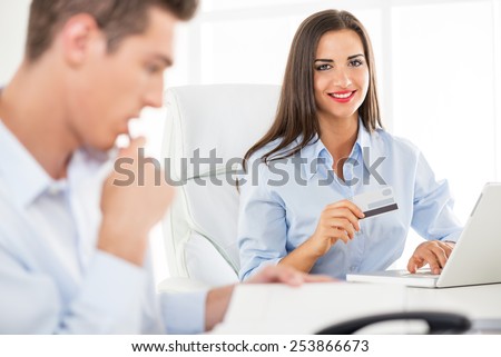 Young business people, woman and man, sitting in an office, a young businesswoman which is in the foreground holding a credit card and with a smile looking at the camera.