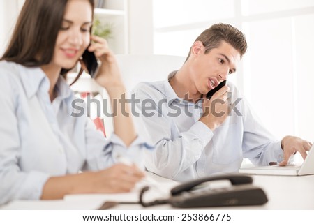 Young business people, man and woman in the office sitting at office desk and make phone calls. The focus is on the young man.