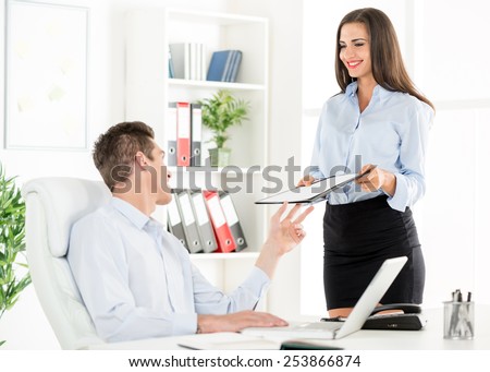 Young business people in the office, a woman standing next to office desk with a smile on her face gives the folder to young businessman who sits in office chair.