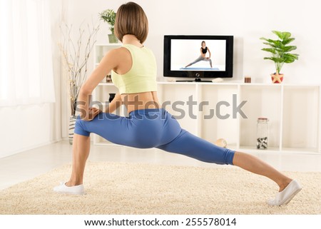 Young woman in sports clothes, athletic build, photographed from behind, exercise in the room, in front of the TV.