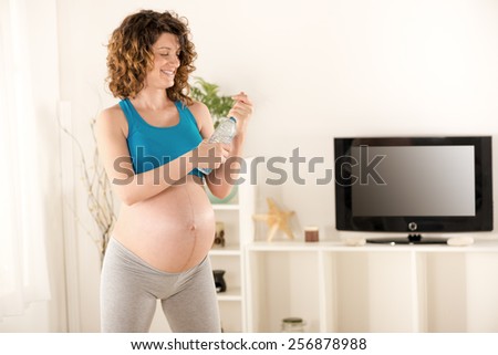 Pregnant woman in sports clothes, standing in the living room, holding a bottle of water in hand and opens it with a smile on her face.