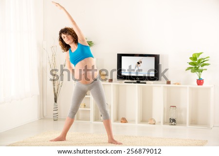 Pregnant woman in sports clothes doing the exercises in the living room, in the background you can see the TV on which is displayed exercises.