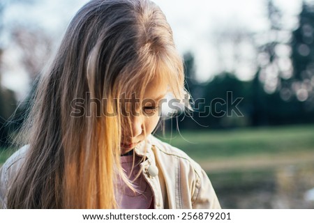 Close-up of a little girl with long hair, standing alone, head bowed, looking down with a sad expression on her face.