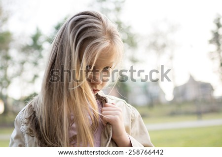 Close-up of a little girl with long hair standing alone outside with a sad expression on her face looking at the camera.