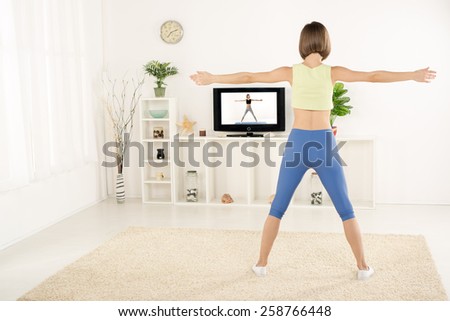 Young woman in sports clothes, photographed from behind, exercise in the room, watching the exercises on TV.