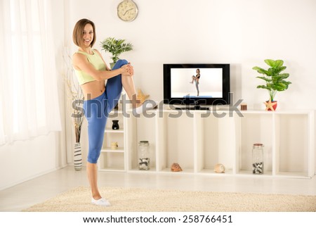 Young woman in sports clothes, athletic build,  exercise in the room, in front of the TV with a smile looking at the camera.