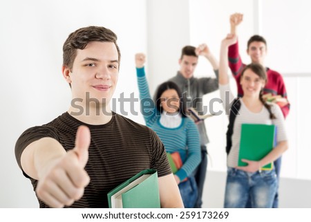 A beautiful smiling young man with book and thumb up is standing in the foreground. A happy group of his friends is behind him with arms in the air.