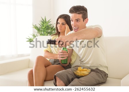 Young man sitting on sofa and watching football on tv. His girlfriend is behind, frowning because and bored.