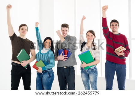 Group of cheerful students with books standing with arms raised in a fist in school hall and looking at camera.