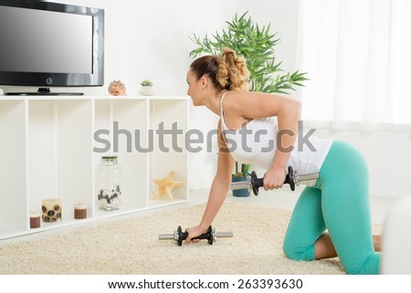 Beautiful young woman doing exercise to strengthen backs with dumbbells at home in front of the tv.
