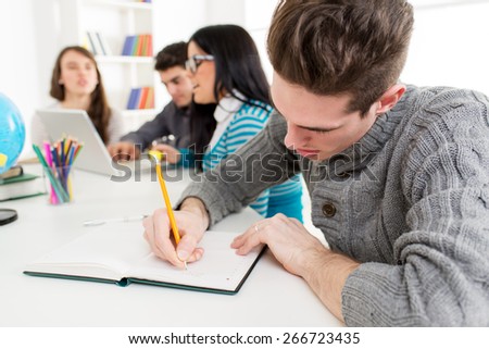 Young man student sitting and writing in the foreground. A happy group of his friends is behind him and looking at laptop.
