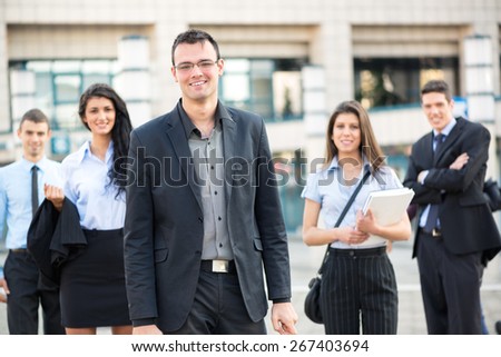 Young businessman, elegantly dressed, standing proudly with his team front of office building.