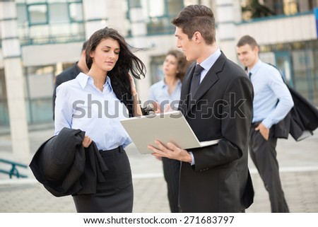 Young businessman talking to his business partner in front of office building holding a laptop while in the background see the rest of the business team.