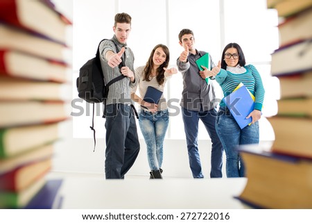 Group of cheerful students standing among the book\'s with thumb up in school hall and looking at camera.